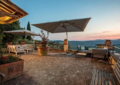 Large stone terrace with parasols, lounge seating and potted plants overlooking the Tuscan hills at Villa Erbolino