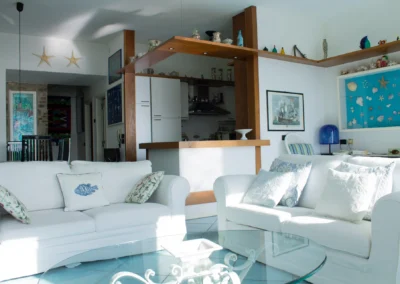 Open-plan living area with white sofas, a glass coffee table and the kitchen behind, decorated with shells and sea art