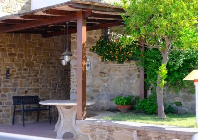 Wooden pergola against a stone wall with a stone table and orange tree, by the outdoor kitchen